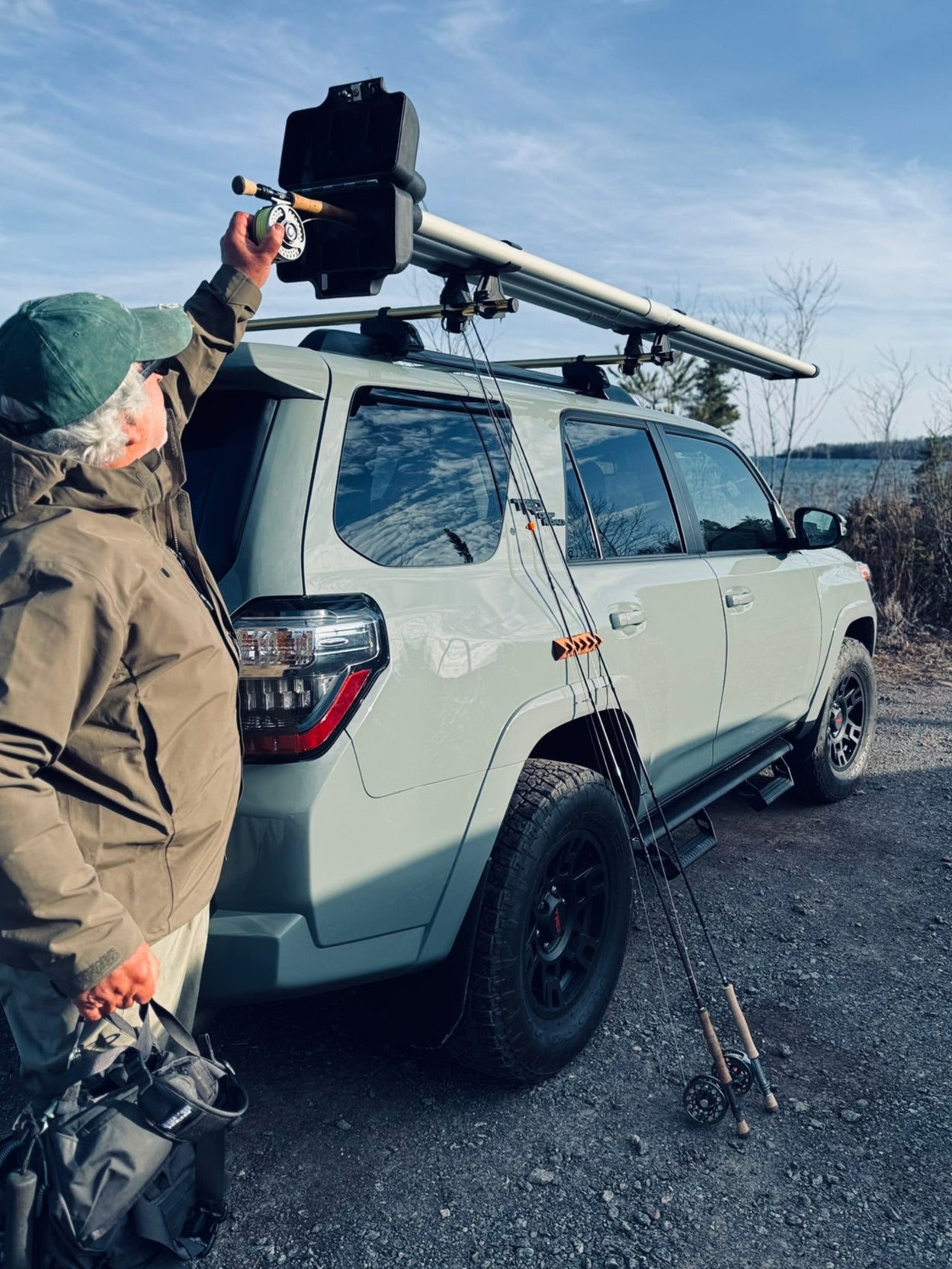 Fly rods secured upright to the fender of a cement colored SUV using the CastHitch magnetic fly rod holder, parked riverside lakeside with a man taking a flyrod off the top of the vehicle.