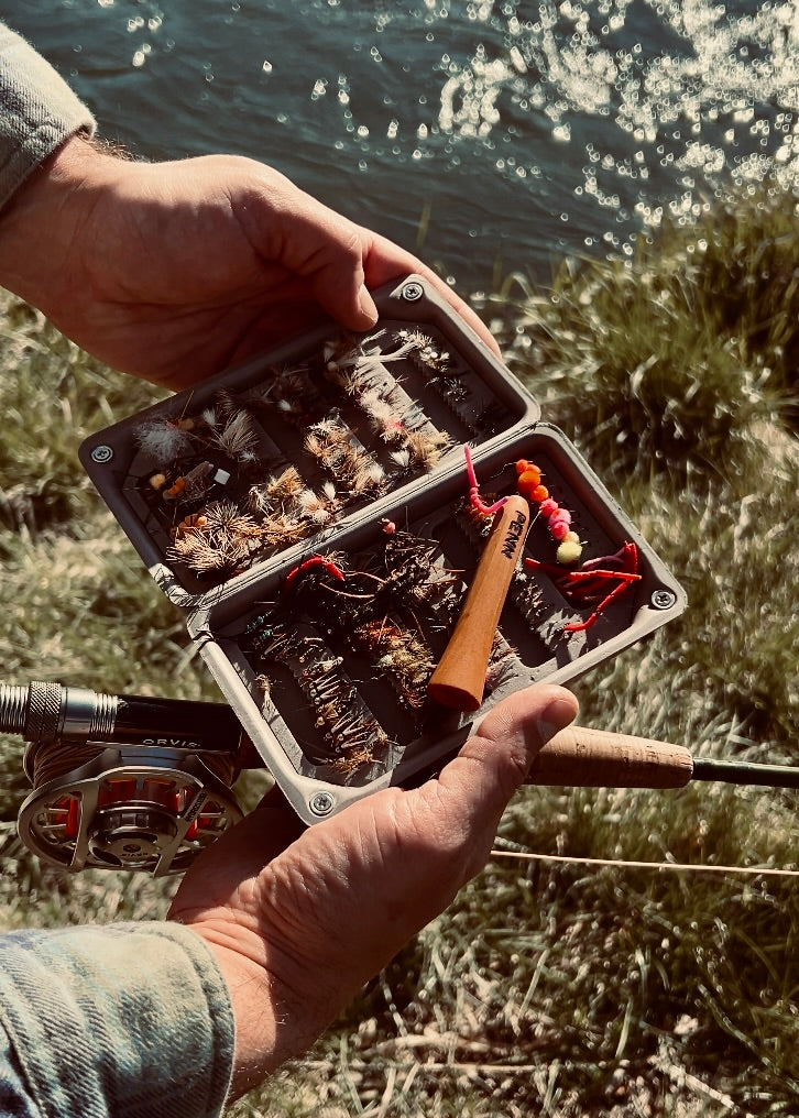Angler holding an open fly box filled with hand-tied flies, a Penn One-Note Fish Whistle resting inside, and a fly rod in hand. Sunlight glistens off the river in the background, capturing the essence of a laid-back day on the water—perfect for casting, unwinding, and embracing the ritual of fly fishing with a cold beer or a well-earned smoke.