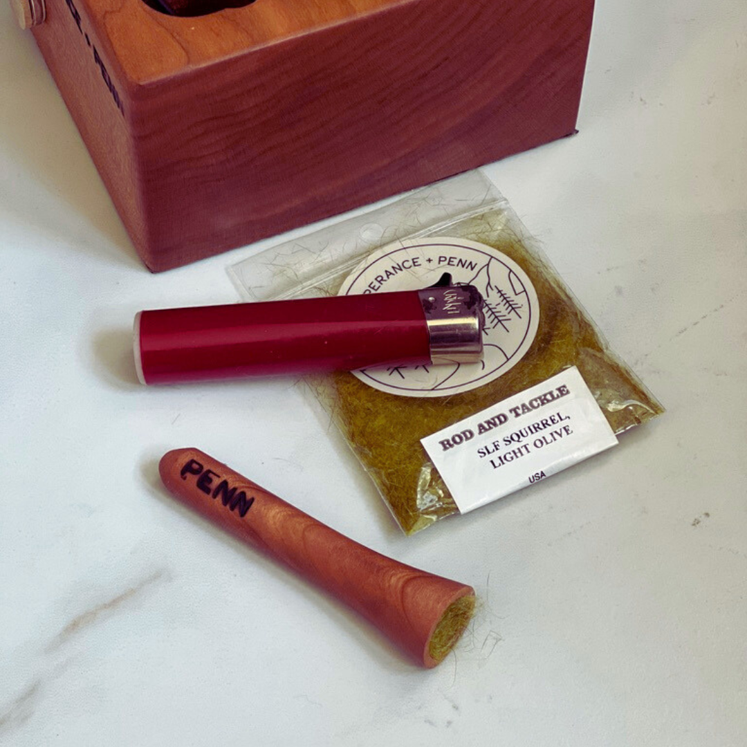 The Penn One-Note Fish Whistle, a handcrafted wooden smoking pipe for fly fishing anglers and cannabis enthusiasts, resting beside a red lighter and a pack of dubbing material labeled 'Rod and Tackle SLF Squirrel Light Olive.' A wooden fly tying caddy from Temperance + Penn sits in the background, blending fly fishing tradition with 420-friendly culture.